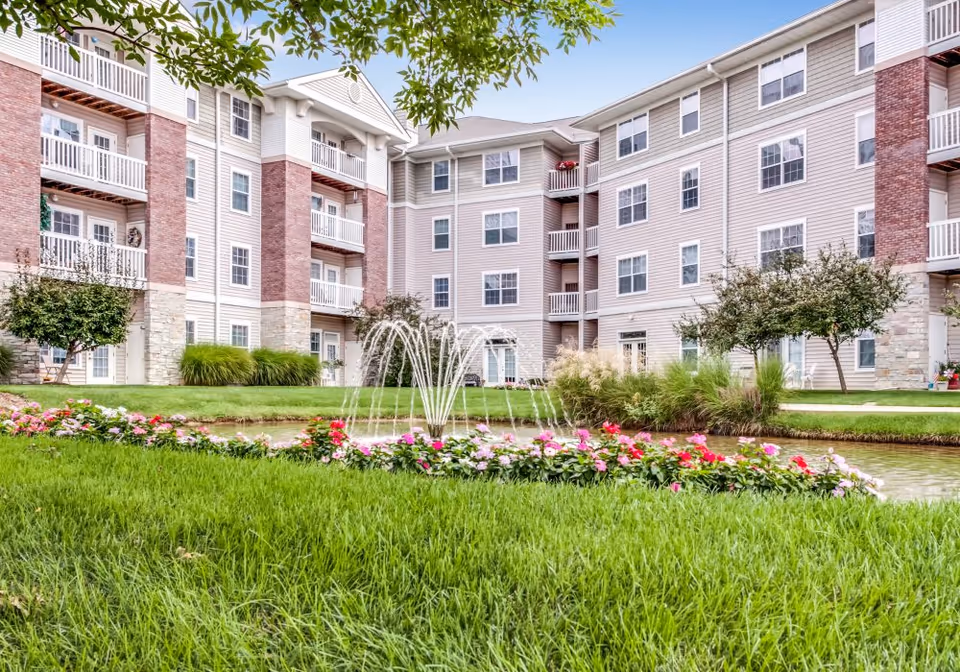 Exterior view of a multi-story senior living facility building with balconies, surrounded by well-maintained green grass, flowering plants, small trees, and a water fountain in the foreground.