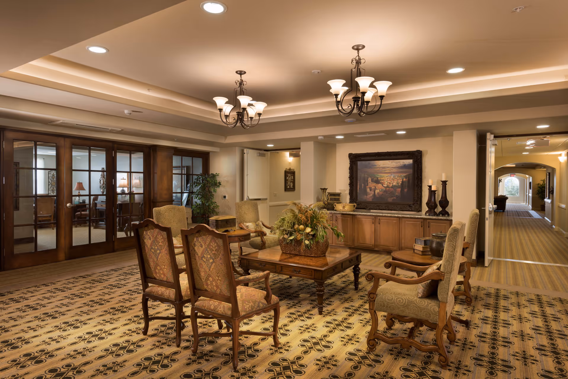 A cozy senior living facility common area with a patterned carpet, wooden chairs with upholstered seats arranged around a wooden coffee table with a floral centerpiece. The room features warm lighting from chandeliers, a framed painting on the wall, decorative vases, and a hallway leading to other rooms. Glass-paneled wooden doors are visible on one side.
