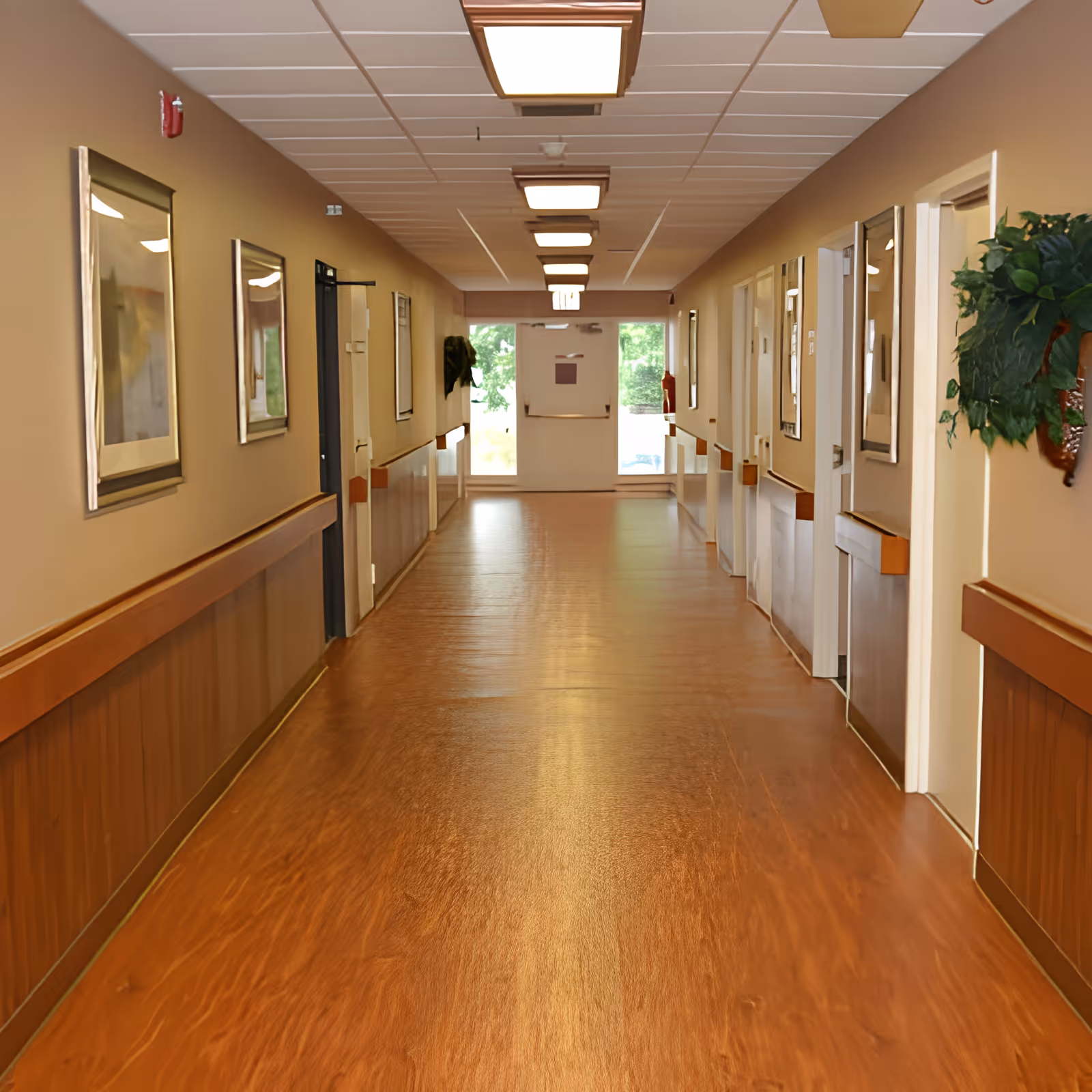 Long, well-lit hallway in a nursing facility with wooden floors, handrails, framed artwork and doors along both sides.