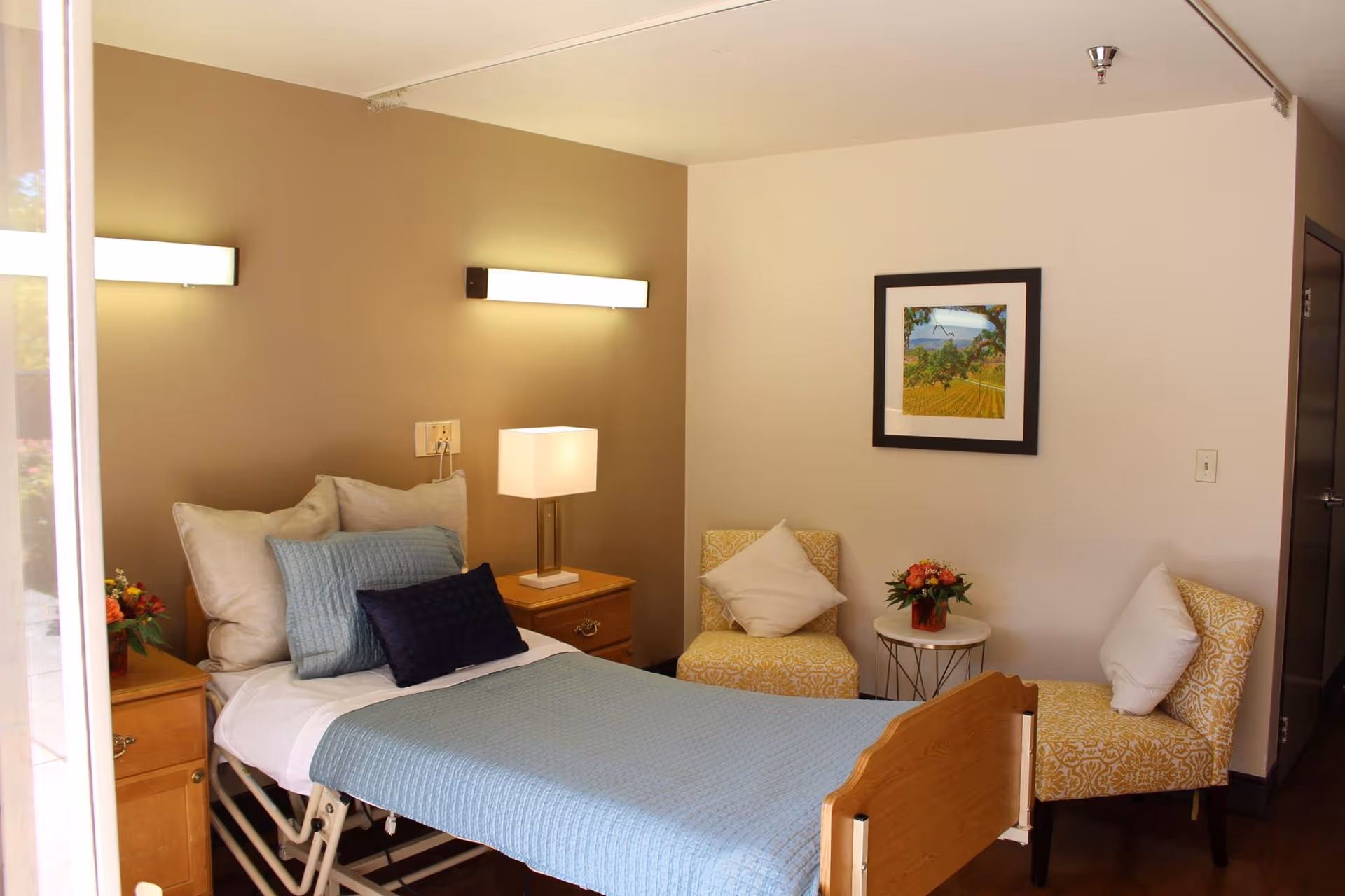 A cozy bedroom in a senior living facility featuring a single adjustable bed with blue bedding and multiple pillows. There are two wooden nightstands on either side of the bed, one with a table lamp and the other with a small flower arrangement. Two patterned armchairs with white pillows are positioned around a small round table with a flower vase. A framed landscape picture hangs on the wall above the chairs. The room has warm lighting and a calm, inviting atmosphere.