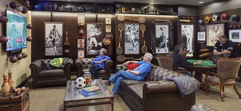 A cozy living room with dark leather sofas and armchairs where three elderly people are seated, watching a sports game on a wall-mounted TV. The walls are decorated with vintage sports memorabilia including framed black-and-white photos, tennis rackets, boxing gloves, and various sports balls. There is also a small table with books and a soccer ball in the center of the room, and two people are playing cards at a table in the background.