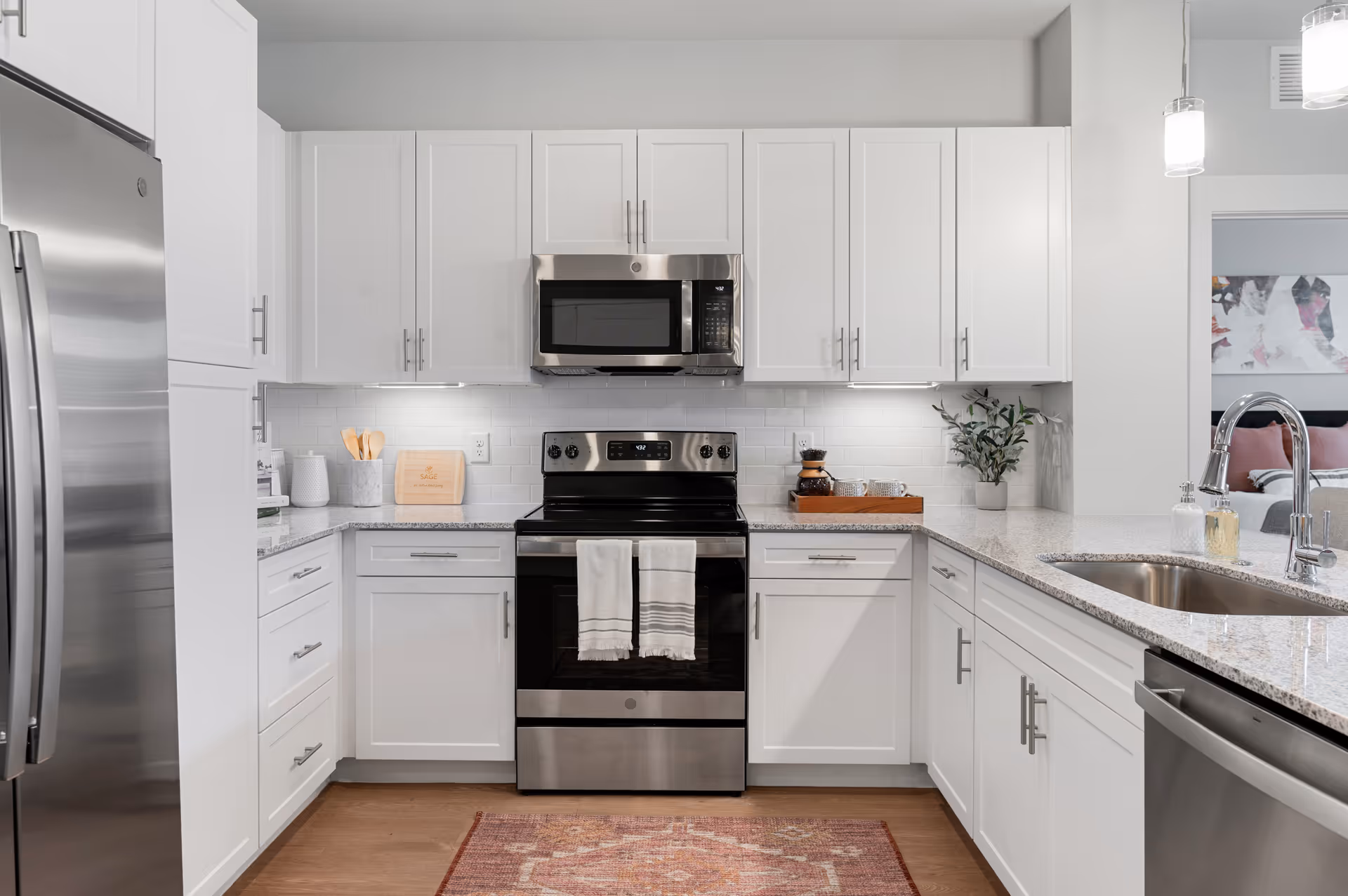 Modern kitchen with white cabinets, stainless steel refrigerator, stove, microwave, and dishwasher. Granite countertops with a sink and faucet on the right side. Two white towels hang on the oven handle. A small plant and a tray with cups and a coffee pot are on the countertop. A wooden cutting board with the word 'SAGE' is visible on the left side. A patterned rug is on the wooden floor in front of the stove.