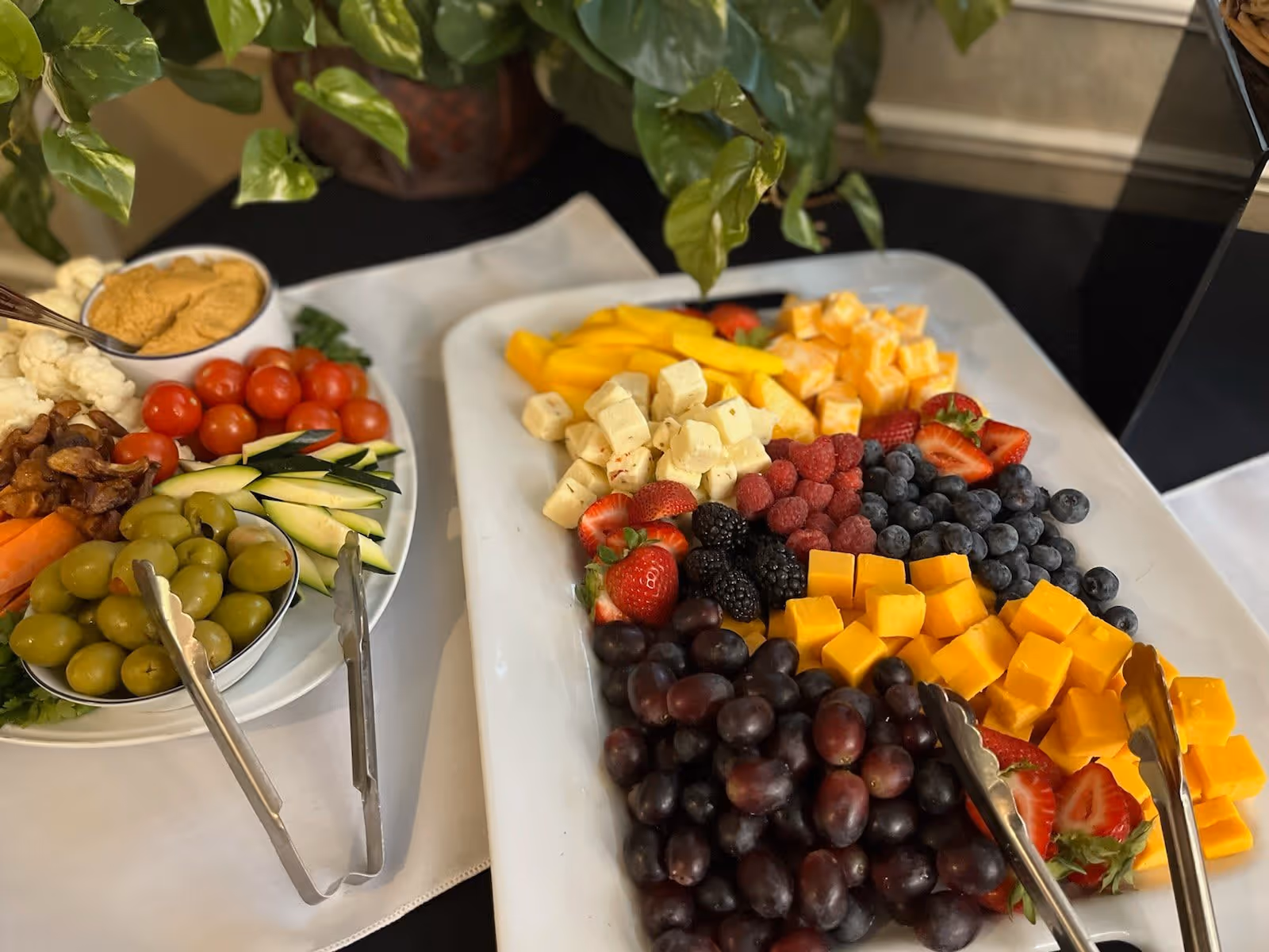 A close-up view of a food platter featuring an assortment of fresh fruits including strawberries, blackberries, raspberries, blueberries, grapes, and cubed cheese. Next to it is another plate with green olives, cherry tomatoes, sliced cucumbers, cauliflower, carrots, and a bowl of dip. Metal tongs are placed on both plates, and a green leafy plant is visible in the background.