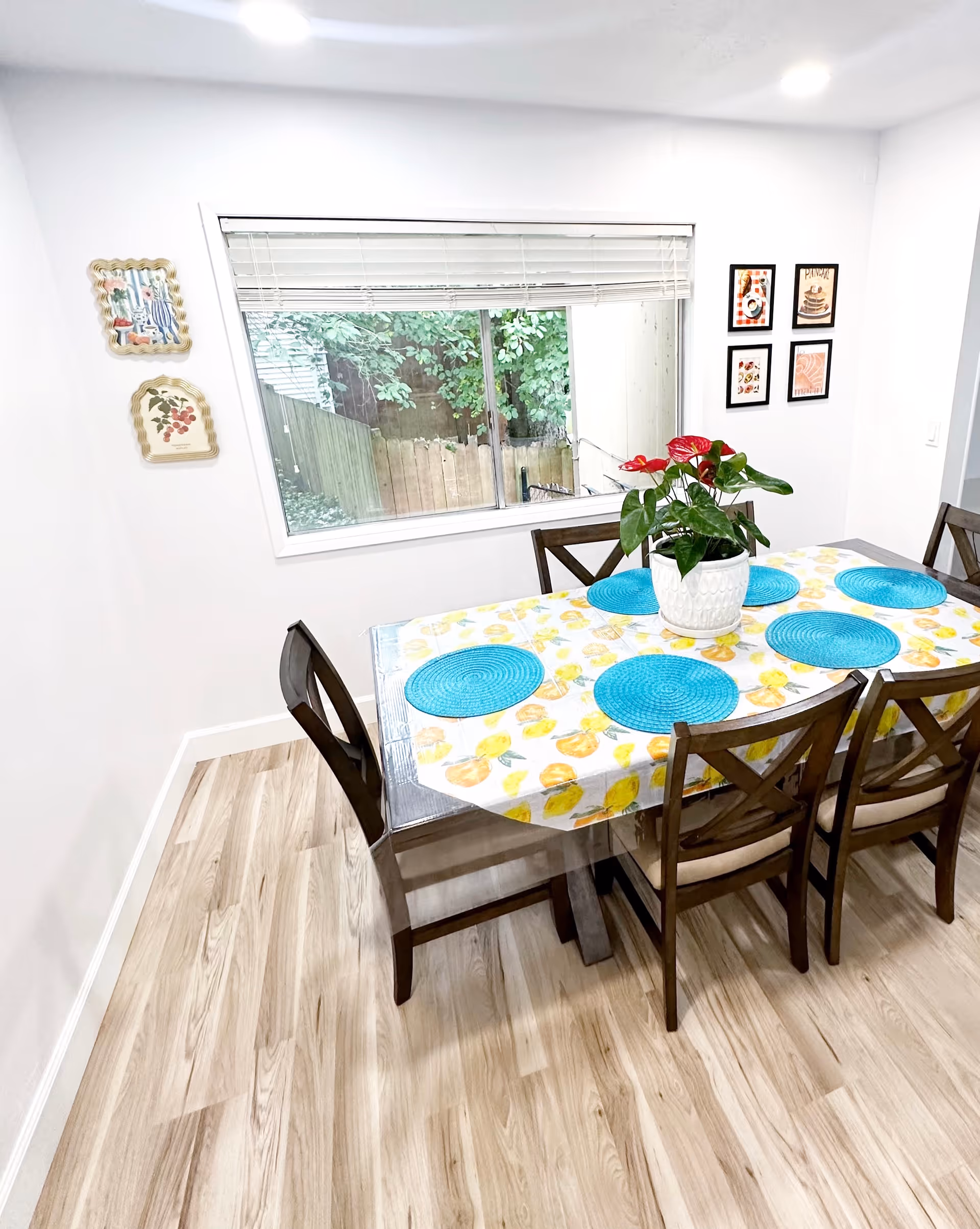 Dining room with a wooden table set with blue placemats and a potted plant beneath a large window.