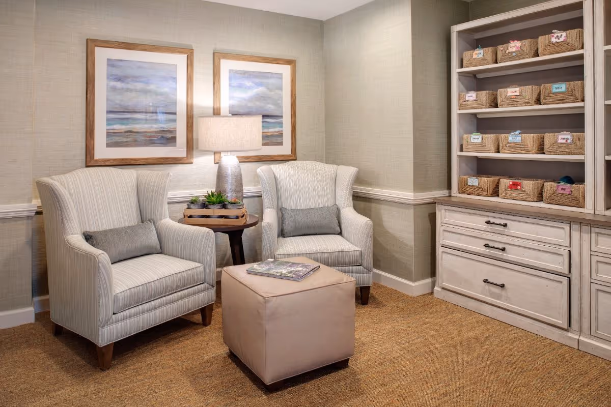 A cozy sitting area with two striped armchairs, a beige ottoman with a book on top, a small round wooden table with a lamp and decorative plants, and a white shelving unit filled with labeled wicker baskets. Two framed ocean-themed paintings hang on the wall behind the chairs.