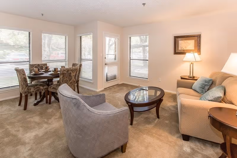 A cozy living room area with a beige sofa adorned with blue cushions, a wooden coffee table with a glass top, and a gray upholstered armchair. To the left, there is a small round dining table with four patterned chairs set near three large windows with blinds. A door with a window is also visible, letting in natural light. The room has beige carpet and walls, with a framed picture and two table lamps providing warm lighting.
