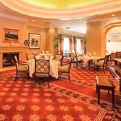 Warm, elegant dining room with round tables and upholstered chairs, a fireplace, and ornate carpeting.