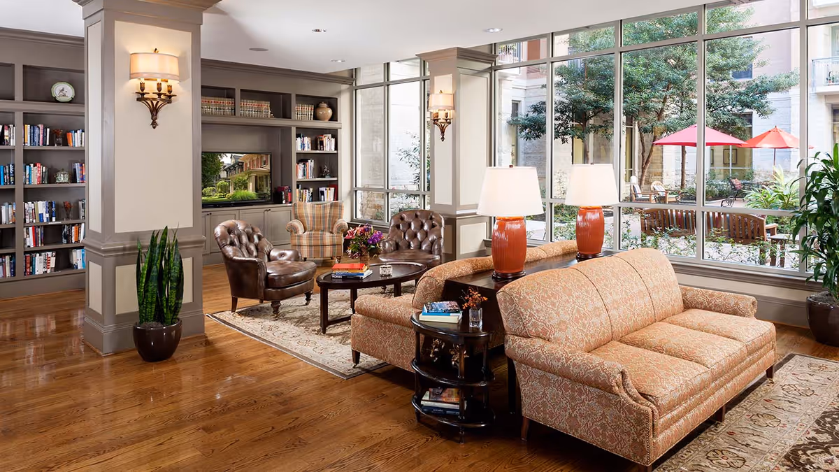 Well-lit common lounge with upholstered sofas, leather chairs, bookshelves, a coffee table and large windows overlooking a courtyard with red umbrellas.