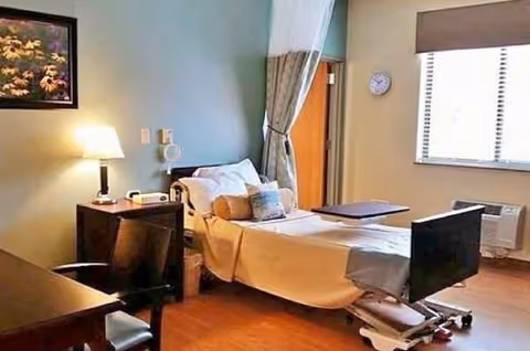 Hospital-style patient bedroom with a bed, overbed table, desk, lamp, curtains, and window.