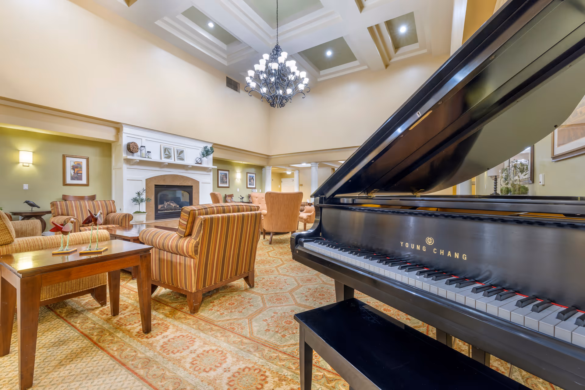 A spacious and elegant living room area in a senior living facility featuring a black grand piano in the foreground, several striped and solid upholstered armchairs arranged around a coffee table, a fireplace with decorative items on the mantle, framed artwork on the walls, and a large chandelier hanging from a coffered ceiling.