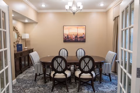 A dining room with a dark wooden oval table surrounded by six chairs, four with dark wooden frames and two with striped upholstery. The room has beige walls, a patterned carpet, a chandelier hanging from the ceiling, and a framed artwork on the wall. There is a wooden sideboard with a lamp and decorative mirror on the left side, and French doors partially open on the right.