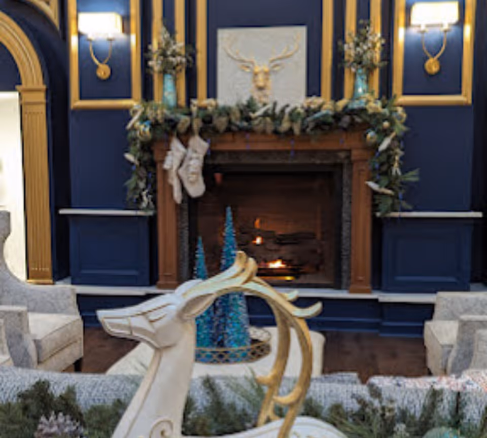 A cozy living room area with a lit fireplace decorated with a garland and hanging stockings. Above the fireplace is a wall-mounted decorative piece featuring a deer head. The walls are painted dark blue with gold trim, and there are two wall sconces with lamps on either side of the fireplace. In front of the fireplace is a white and gold decorative reindeer figure and two small blue Christmas trees. There are upholstered armchairs on either side of the fireplace.