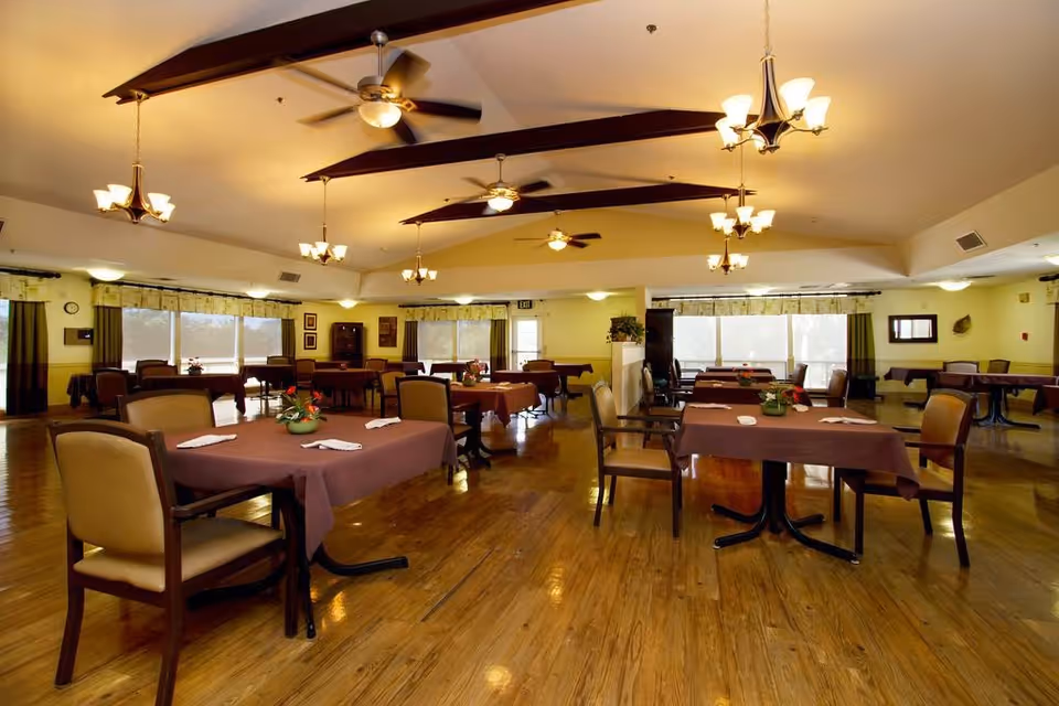 A spacious dining room with multiple tables covered in brown tablecloths, each set with napkins and small flower arrangements. The room has wooden floors, ceiling fans, and hanging light fixtures. Large windows with curtains allow natural light to fill the space.
