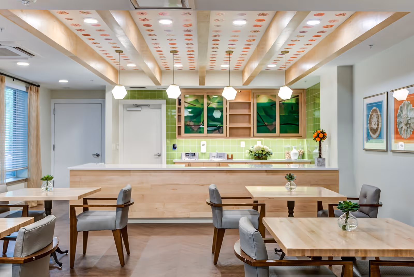 A bright and modern dining area with light wood tables and gray cushioned chairs. The ceiling features wooden beams with decorative patterns, and three pendant lights hang above a light wood counter. Behind the counter, there are green tiled walls and cabinets with glass doors displaying green artwork. The room has large windows with beige curtains and framed floral artwork on the walls.