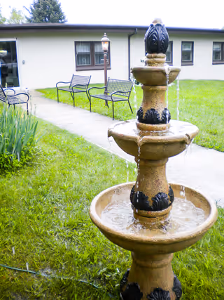 Outdoor area of a senior living facility with a three-tiered stone water fountain in the foreground, green grass, a concrete walkway, two black metal benches, a lamp post, and a single-story building with windows in the background.