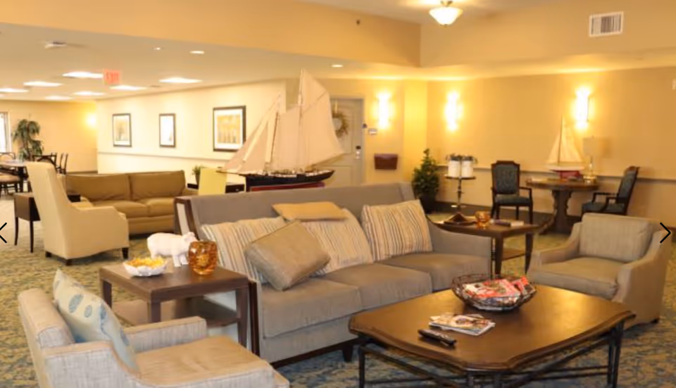 A cozy living room area in a senior living facility with beige walls and carpeted floor. The room features a gray sofa with striped cushions, two matching armchairs, a wooden coffee table with magazines and a basket, and side tables with decorative items. In the background, there are additional seating areas, framed artwork on the walls, and model sailboats as decor.