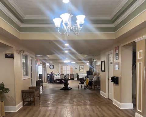 Wide interior hallway and common lounge with a chandelier, seating areas, a central table, and a visible wheelchair/walker.