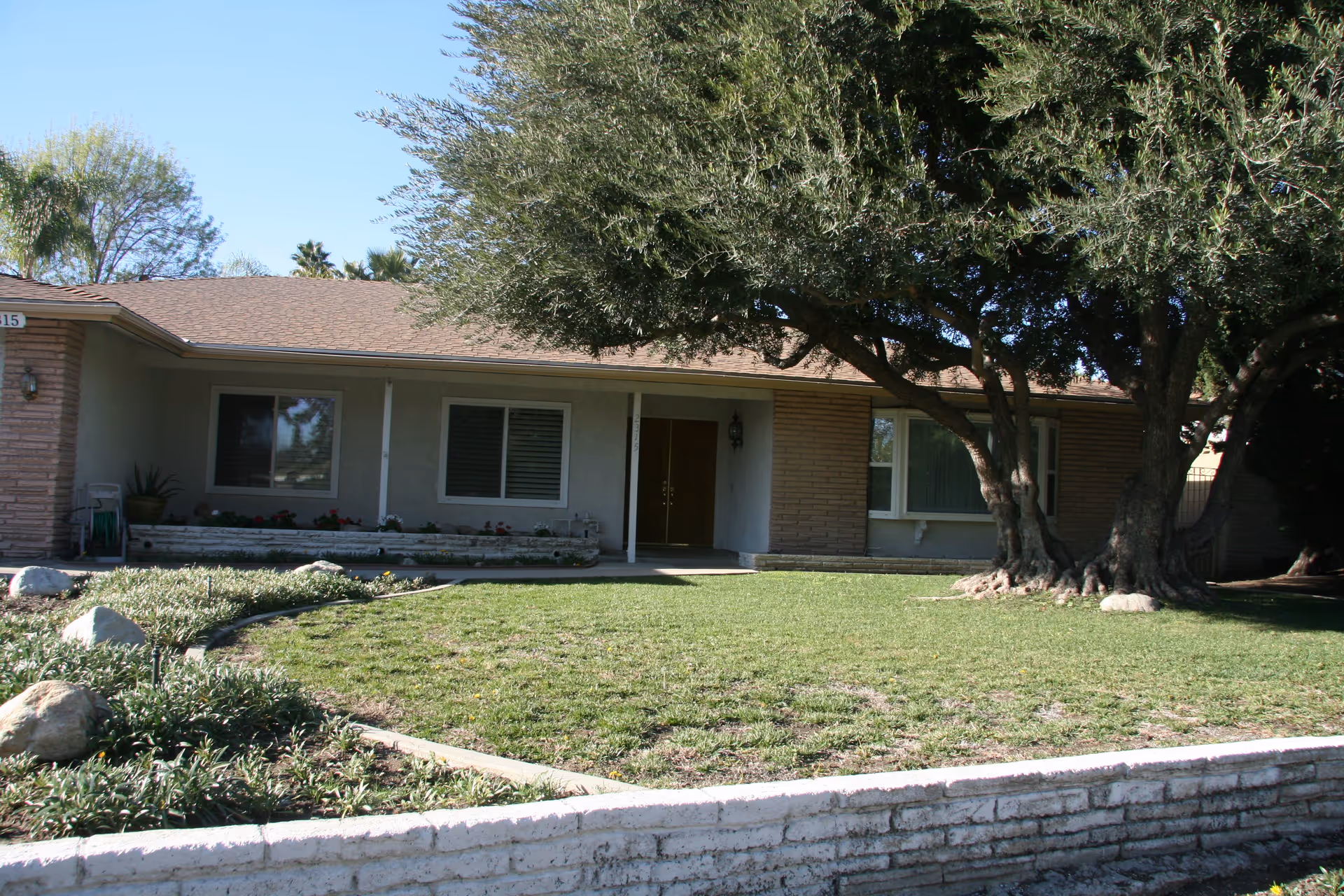 Single-story ranch-style house front with a lawn, large tree, and covered porch entrance.