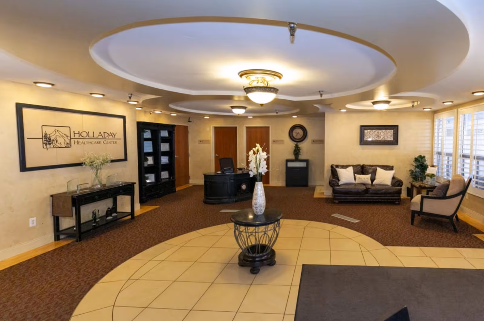 Reception area of Holladay Healthcare Center featuring a circular ceiling design with multiple light fixtures, a central round table with a vase of flowers, a black reception desk, a seating area with a leather couch and chairs, and large windows with white shutters.