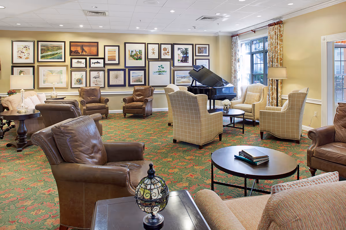 A cozy living room area with multiple armchairs and sofas arranged around coffee tables. The room features a green and red patterned carpet, beige walls adorned with a gallery of framed artwork, and a black grand piano near a window with floral curtains. The space is well-lit with table lamps and natural light from the window.