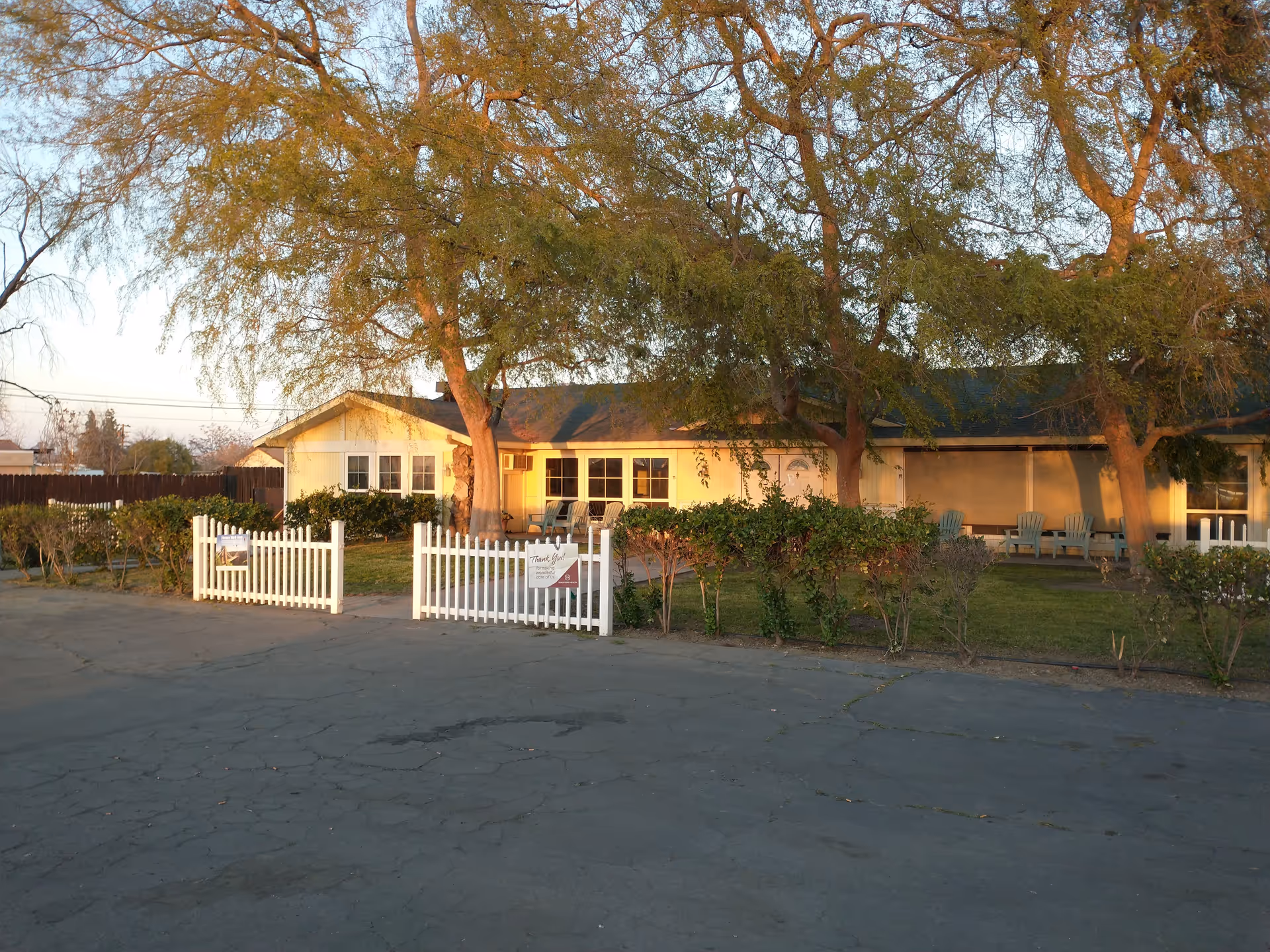 Front exterior of a single-story assisted living building with a white picket fence, lawn, trees, and chairs on the porch.