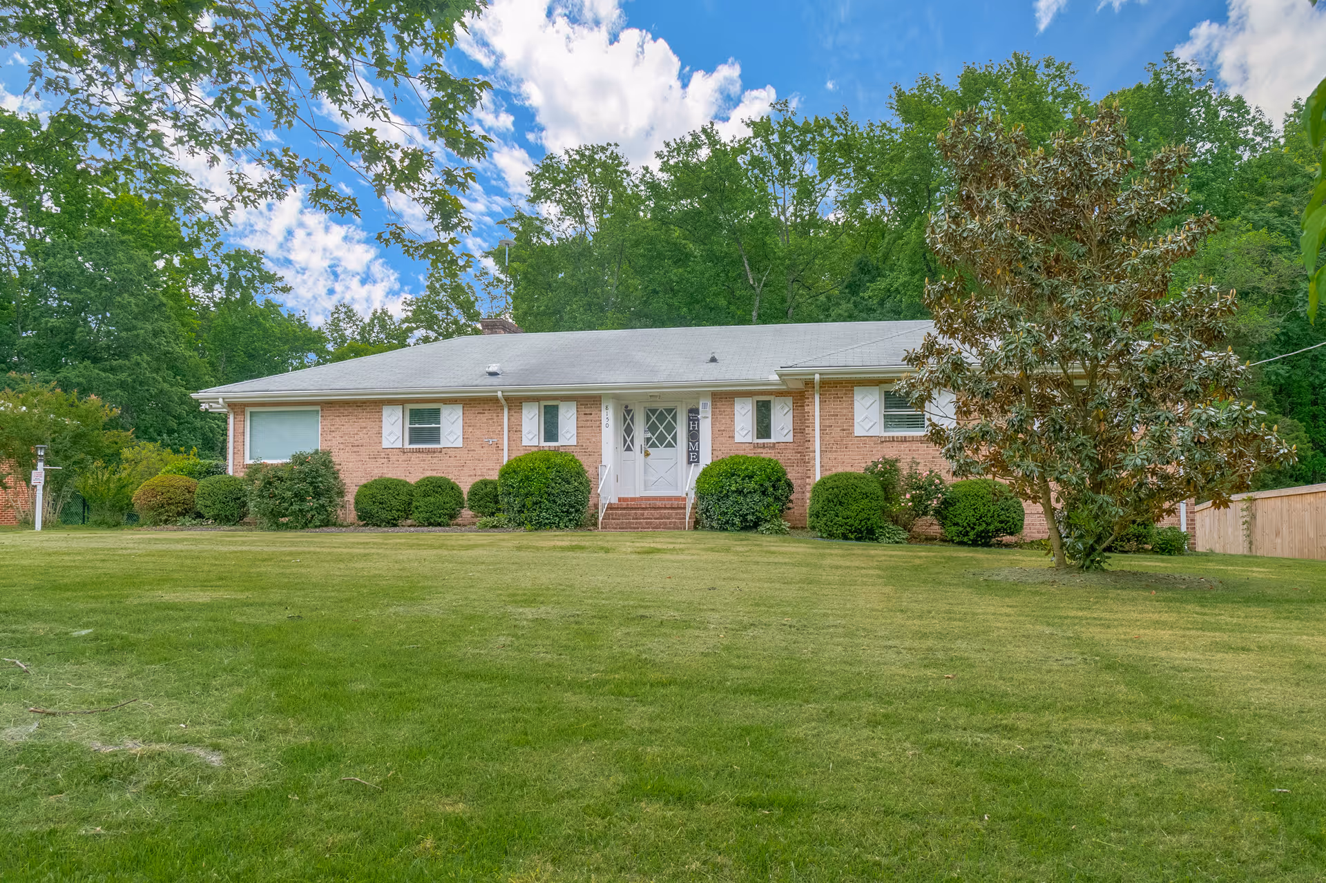A single-story brick house with a gray roof surrounded by green bushes and trees. The house has white window shutters and a white front door with a small set of stairs leading up to it. The lawn in front is well-maintained and green, with a large tree on the right side. The sky is partly cloudy with patches of blue visible.
