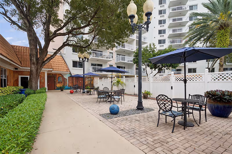 Outdoor patio area with several tables and chairs under blue umbrellas, surrounded by greenery and a white fence. A tall street lamp is in the center, and a multi-story building is visible in the background.
