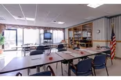 Well-lit interior activity room with rectangular tables and blue chairs arranged classroom-style, a TV, bookshelves, and an American flag near large windows.