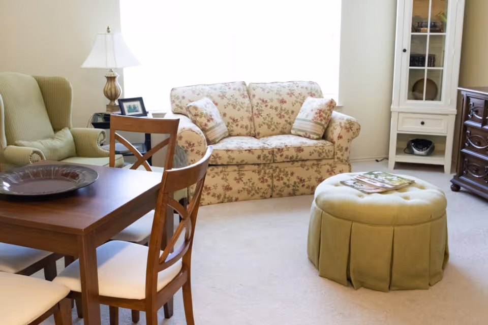 A cozy living room with a floral loveseat, green round ottoman, armchair, and a wooden dining table with chairs.