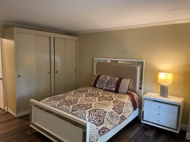 A bedroom in Springfield Senior Living Community featuring a bed with a patterned bedspread and pillows, a white nightstand with a lamp, and a large white wardrobe against a beige wall.