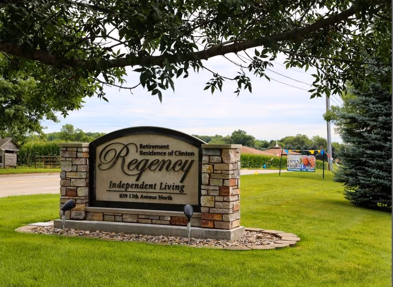 Stone sign for Regency Retirement Residence of Clinton Independent Living located on a grassy area near a road, with trees and a banner advertising an open house in the background.