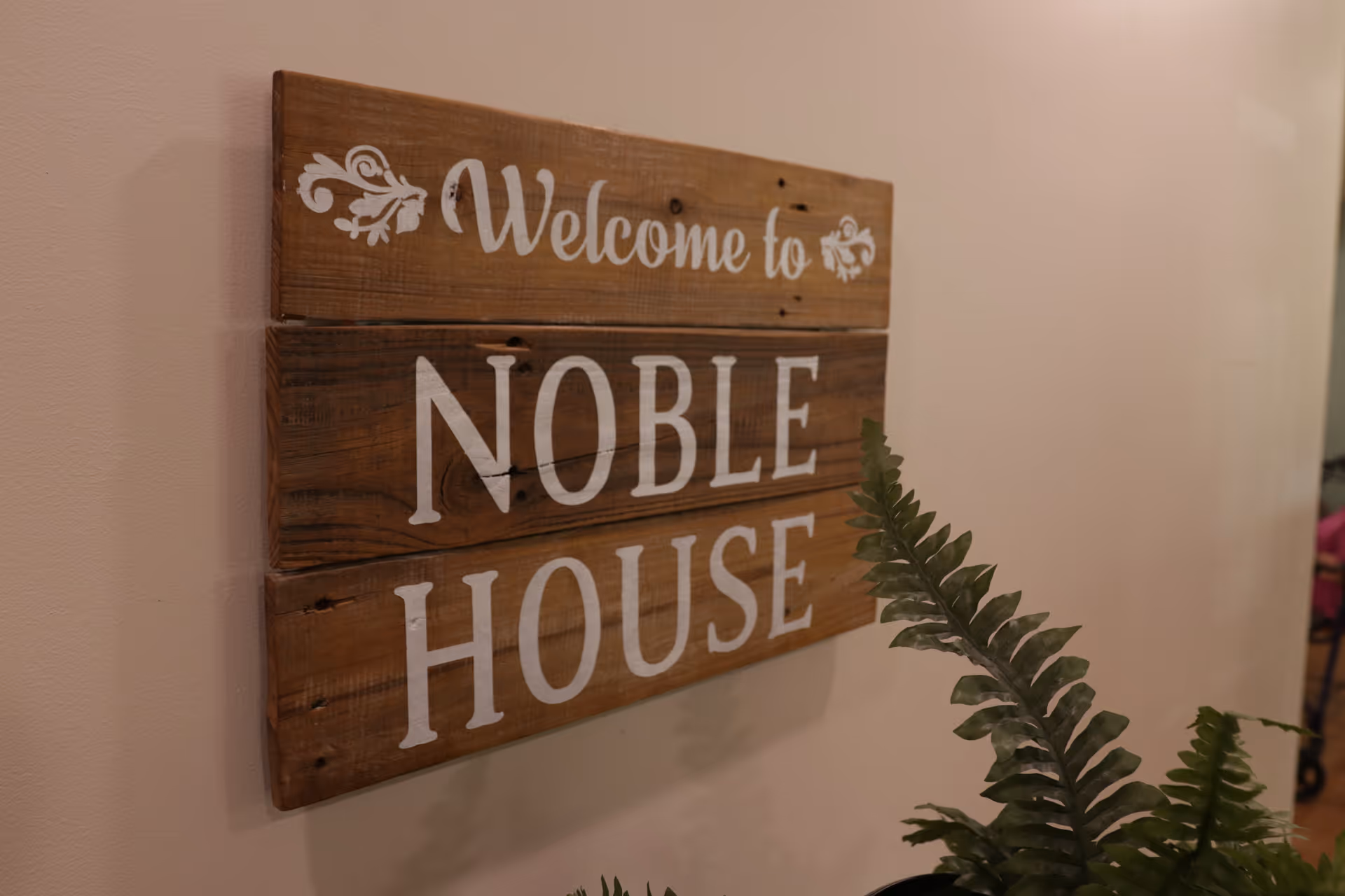 Rustic wooden wall sign reading "Welcome to NOBLE HOUSE" with a potted fern in front.