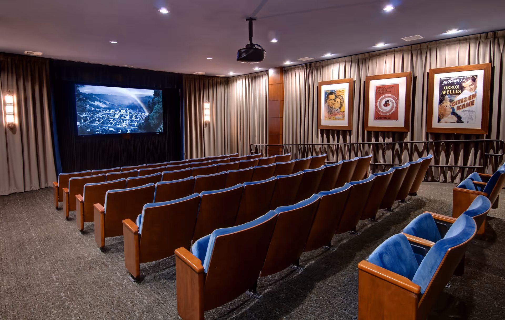 A small movie theater room with rows of wooden seats upholstered in blue fabric facing a large screen showing a black and white movie scene. The walls are lined with beige curtains and three framed classic movie posters are displayed on the right wall. The ceiling has recessed lighting and a projector is mounted in the center.