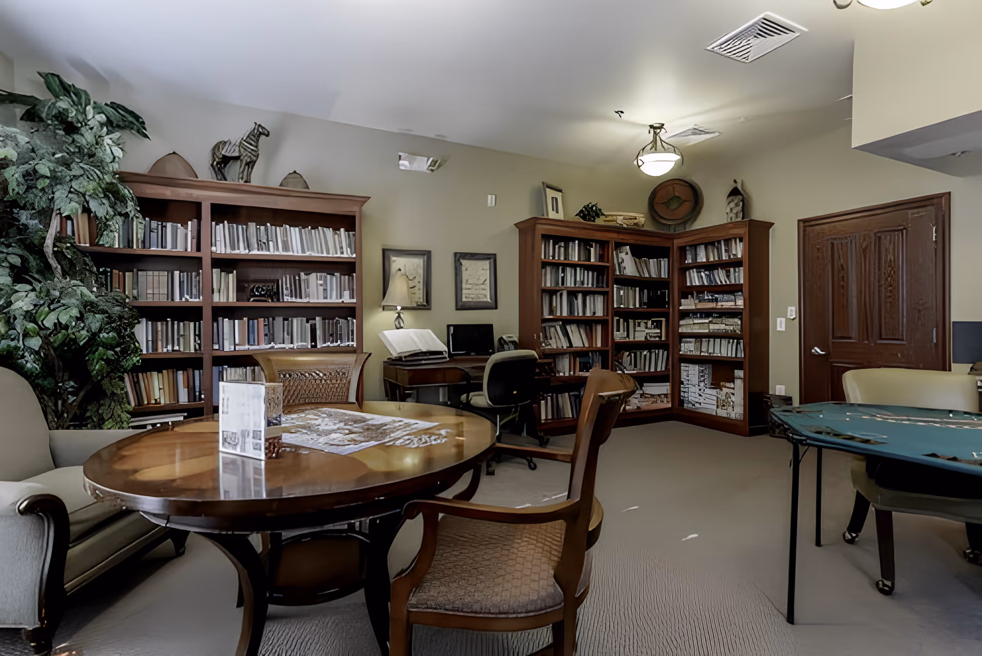 A cozy library or common room in an assisted living facility with wooden bookshelves filled with books, a round wooden table with chairs around it, a computer desk with a chair, framed pictures on the wall, and a green plant in the corner.