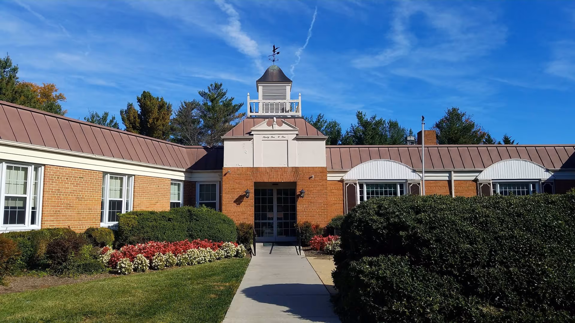 Single-story brick building entrance with a cupola, manicured shrubs and flowerbeds under a blue sky.