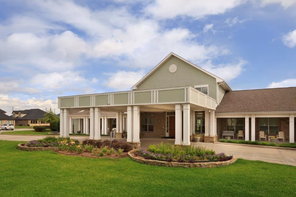 Front exterior view of a single-story assisted living facility with a covered entrance supported by white columns, landscaped flower beds, green lawn, and a partly cloudy sky.