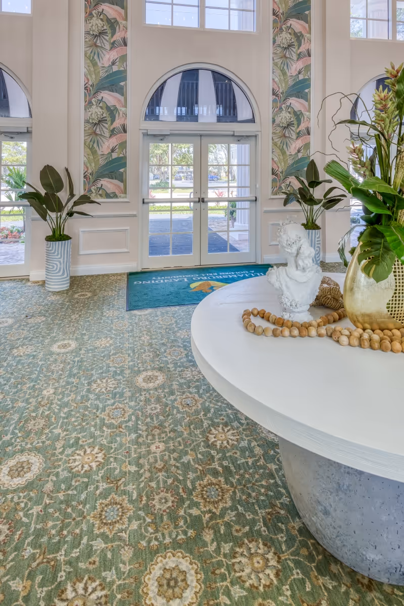 Bright assisted-living lobby with patterned carpet, a round table with decorative items and plants, and double glass entry doors with arched windows.
