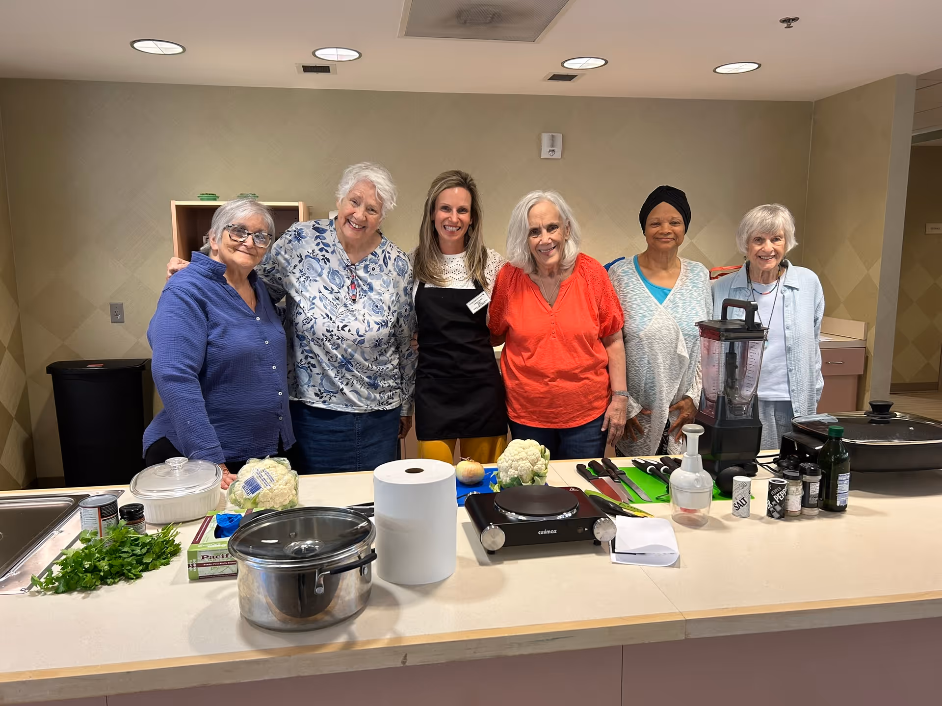 Six women standing behind a kitchen counter with various cooking ingredients and utensils, including a pot, paper towel roll, blender, spices, and vegetables, in a well-lit kitchen area.