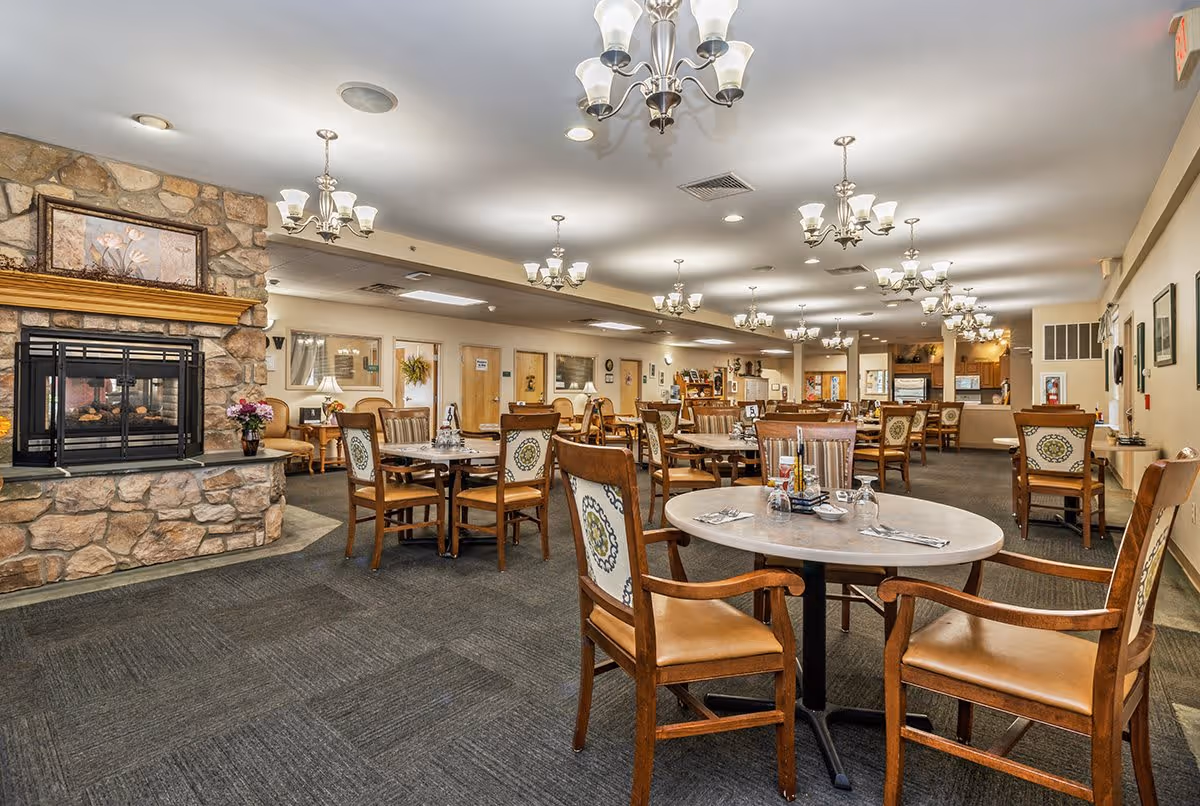 A spacious dining room with multiple round tables and wooden chairs featuring patterned upholstery. The room has a stone fireplace on the left side with a floral painting above it. Several chandeliers hang from the ceiling, providing warm lighting. The walls are decorated with framed pictures and there are additional seating areas visible in the background.