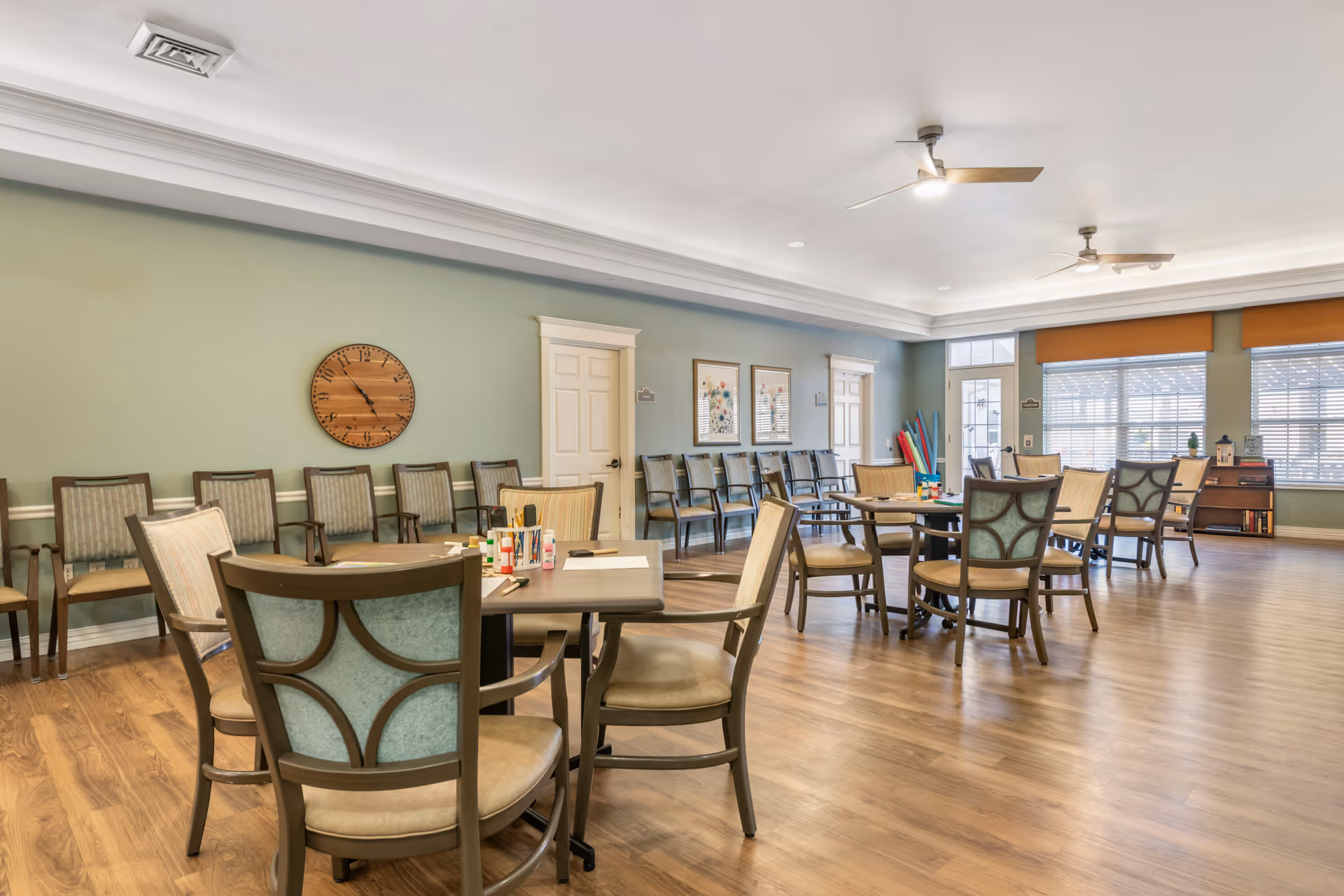 Spacious communal dining/activity room with multiple round tables and chairs, wood floors, large windows, and light green walls.