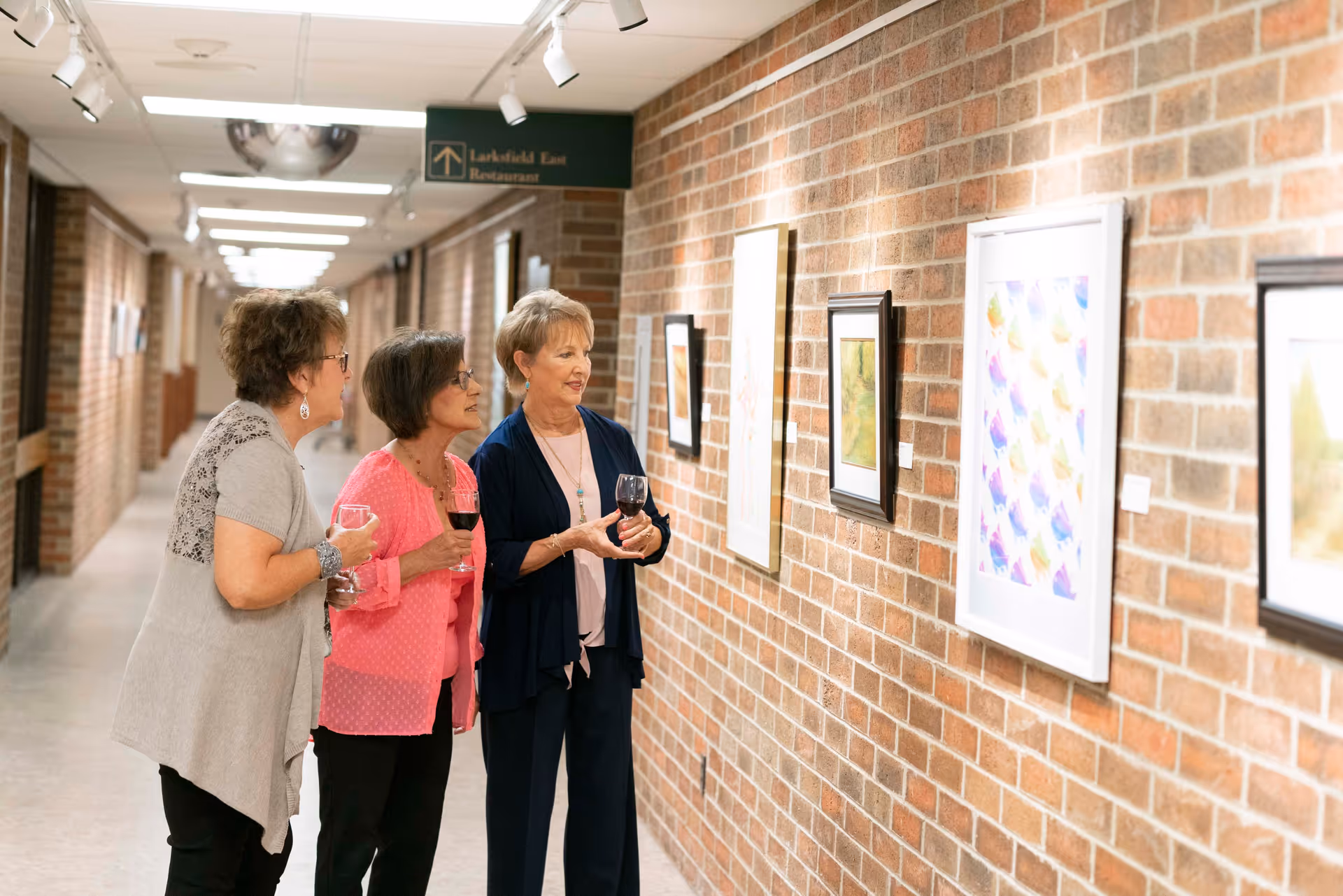 Three elderly women standing in a hallway with brick walls, looking at framed artwork displayed on the wall. They are holding glasses of wine and appear to be engaged in conversation. A sign above indicates the direction to the Larksfield East Restaurant.