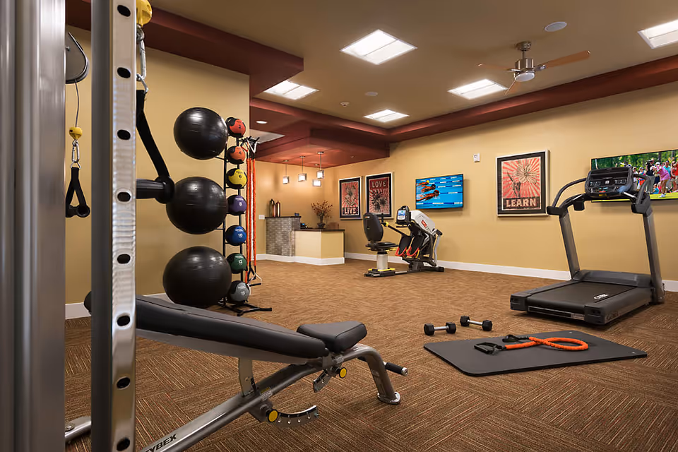 A fitness room with various exercise equipment including a treadmill, stationary bike, weight bench, dumbbells, resistance bands, and large exercise balls. The room has beige walls, brown carpet, ceiling lights, and motivational posters on the walls. There are also two wall-mounted televisions.