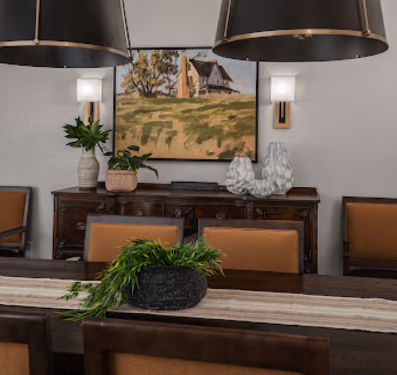 Dining room with a long wooden table, orange-upholstered chairs, pendant lights, plants, and a landscape painting on the wall.