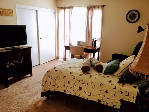 A cozy bedroom with a bed covered in a floral patterned quilt and several pillows. There is a small desk with a chair in front of a window with sheer curtains, a wall clock, a TV on a stand with shelves, and a lamp on a bedside table.
