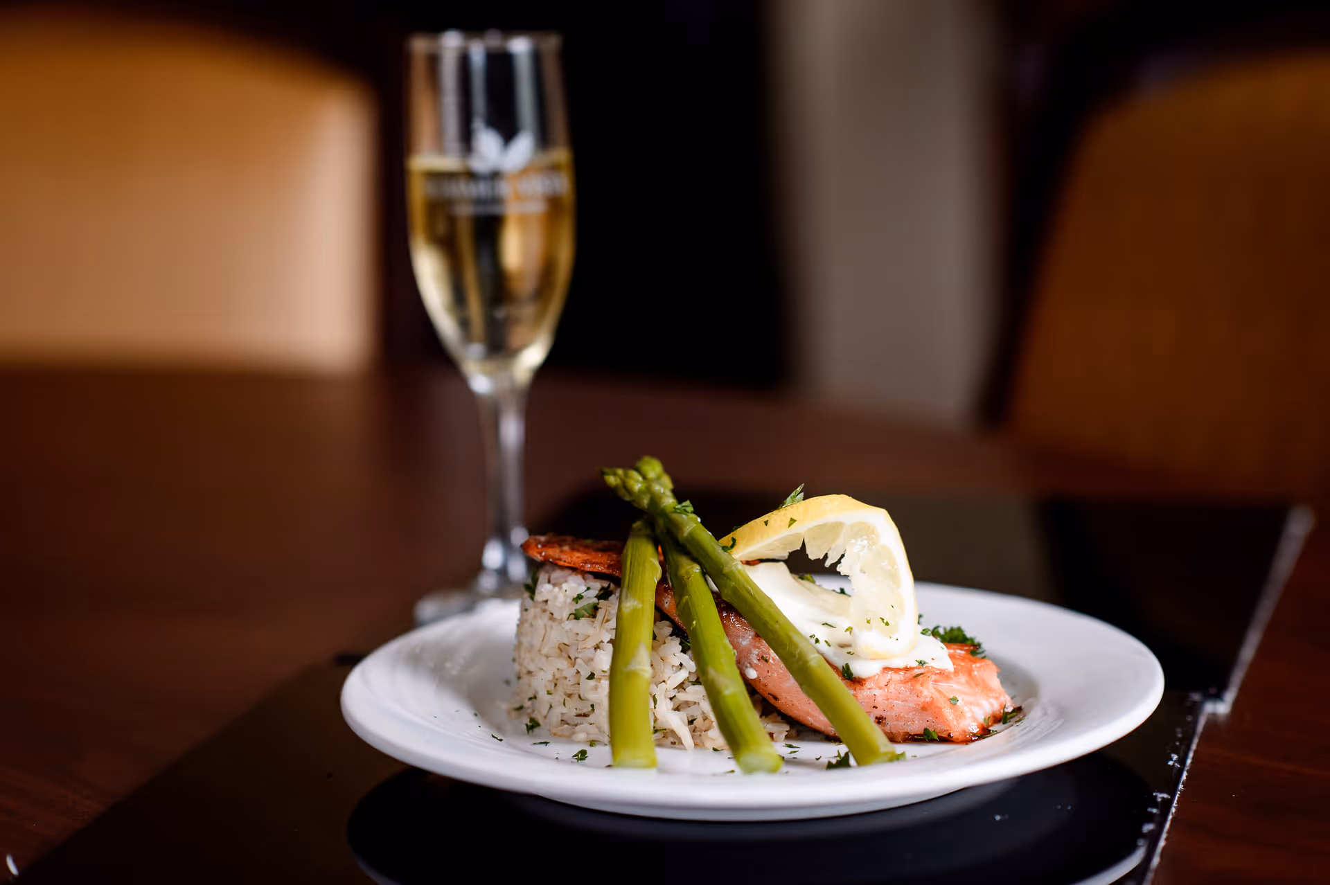 A plated meal consisting of cooked salmon topped with a lemon wedge and three asparagus spears, served with a side of rice. In the background, there is a glass of white wine on a wooden table.