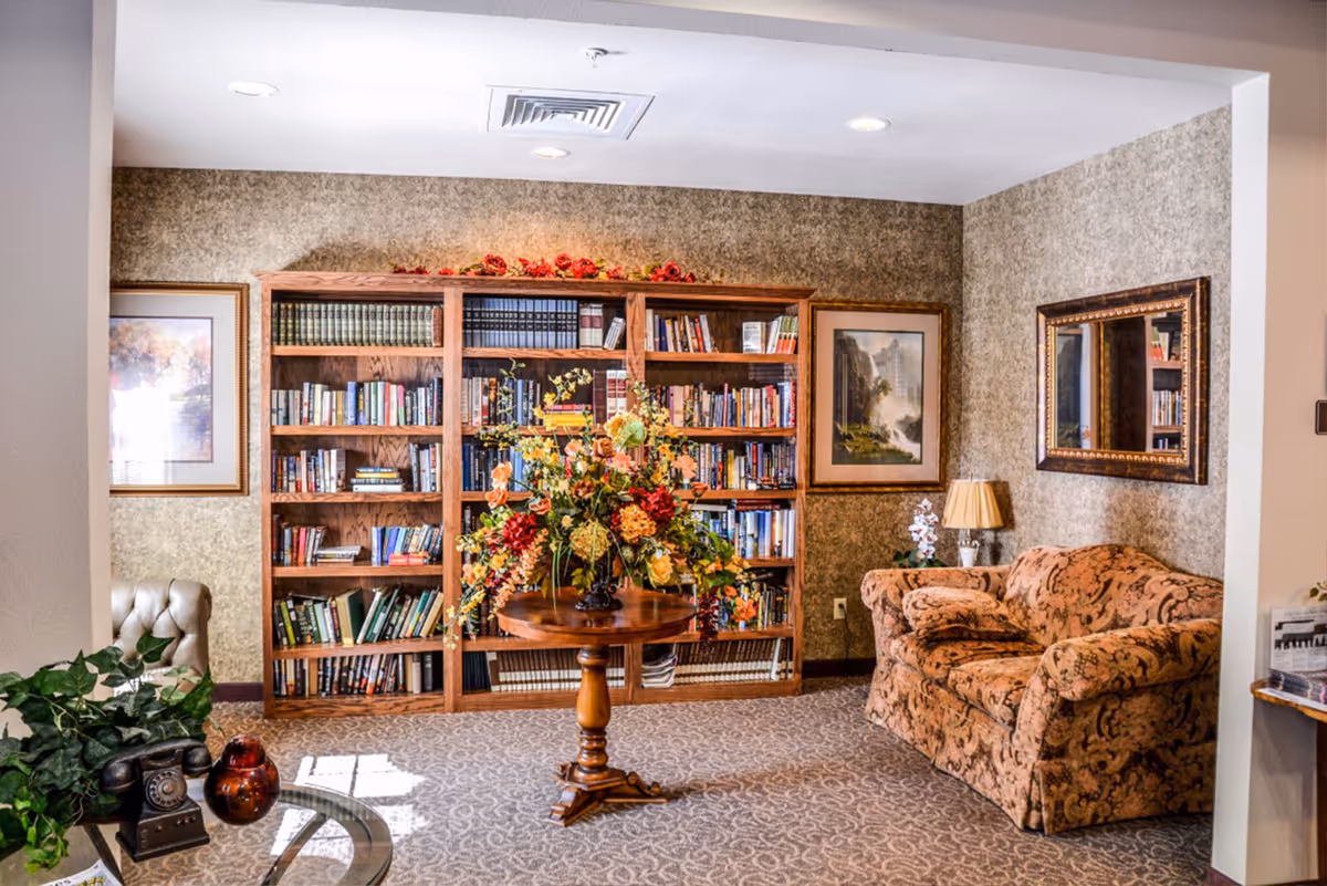 A cozy living room area with a patterned sofa, a wooden bookshelf filled with books, a round wooden table with a large floral arrangement, framed artwork on the walls, a table lamp, and a mirror reflecting part of the room.