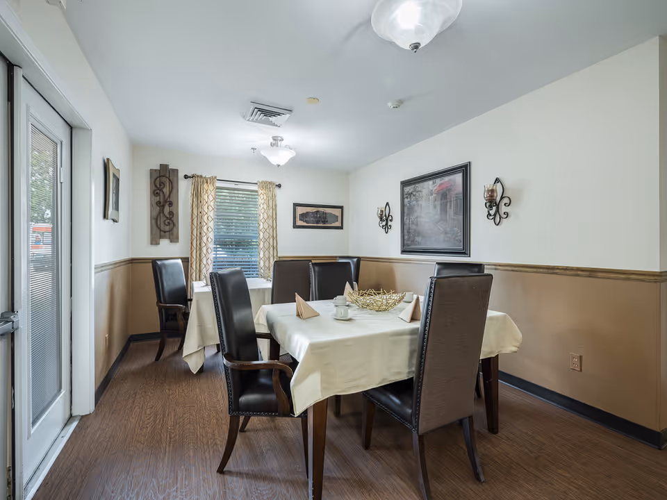 A dining room in an assisted living facility with two tables covered with white tablecloths, each set with folded napkins and cups. The room has wooden flooring, beige and white walls with decorative wall art and sconces, and a window with patterned curtains letting in natural light.