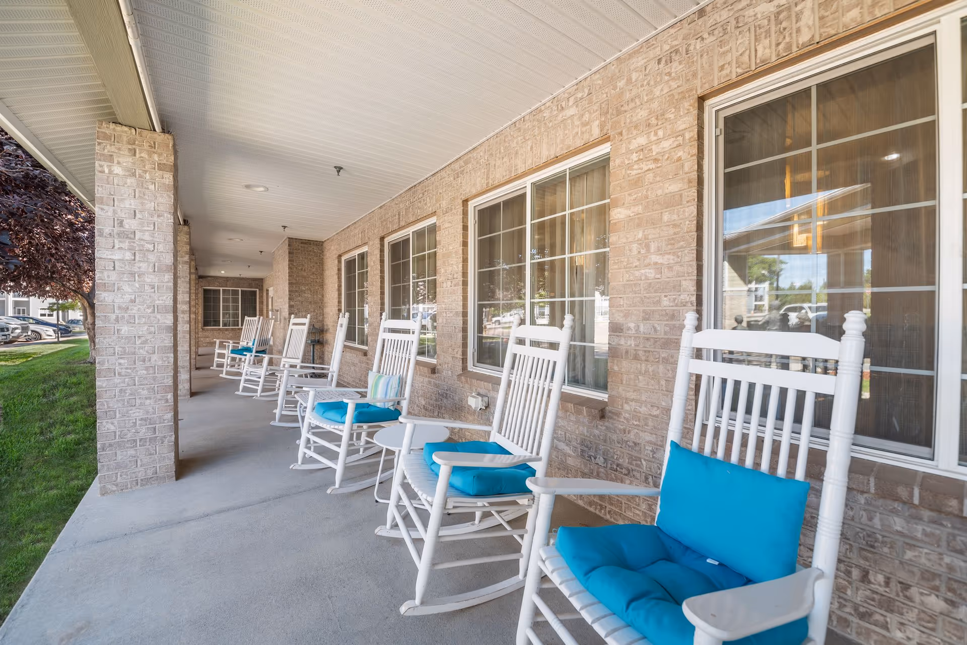 A covered outdoor porch area with a row of white rocking chairs, each with blue cushions and some with additional pillows, positioned along a brick wall with multiple windows. There is a small white side table between some of the chairs, and a grassy area with a tree is visible to the left.