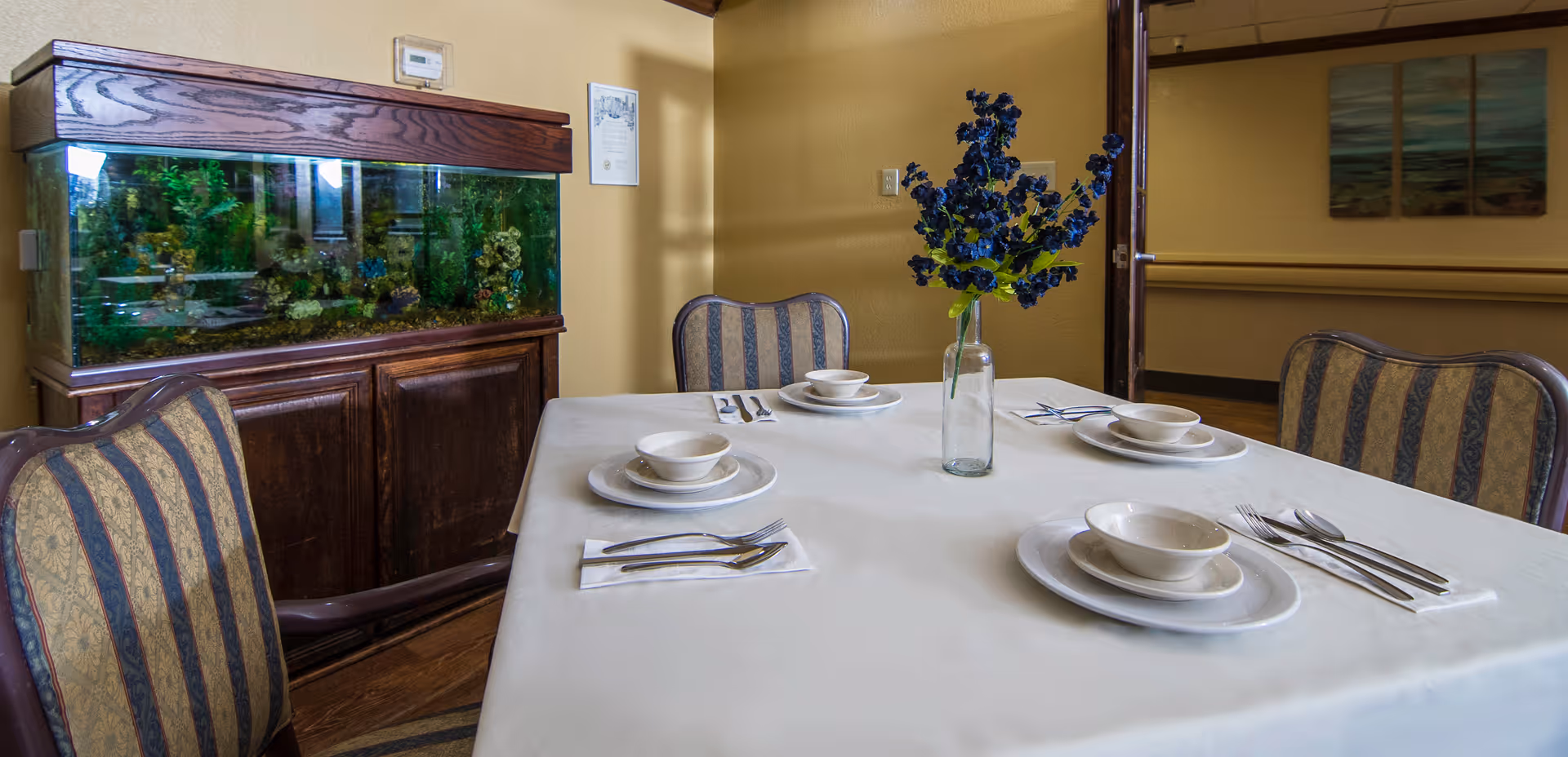 A dining table set for four with white plates, bowls, and silverware on white napkins. A vase with blue flowers is placed in the center of the table. Behind the table is a large fish tank with green plants inside, and the room has beige walls and striped upholstered chairs.