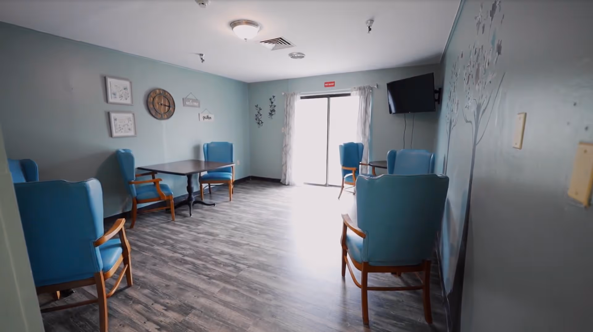 Small communal sitting area with blue armchairs, a table, wall decorations and a wall-mounted TV by a sliding glass door.